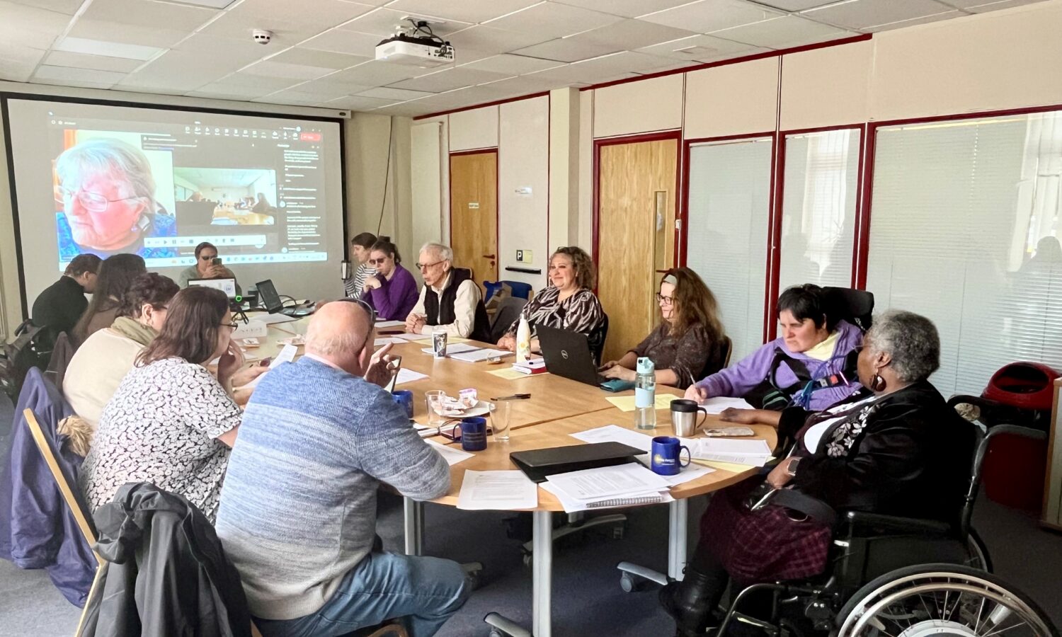 A group sit around a conference table, an attendee can be seen on a screen joining the meeting virtually