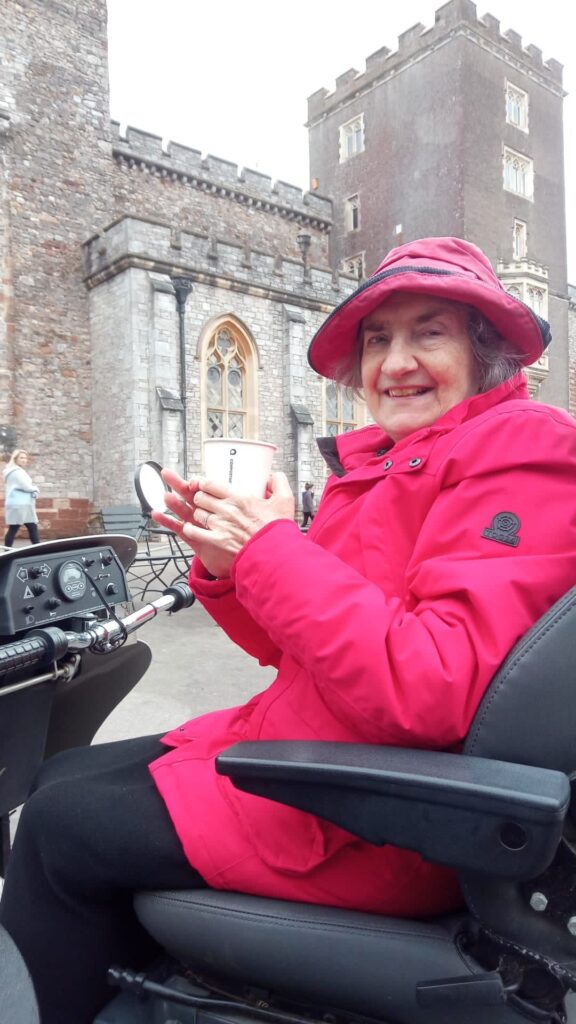 Liz, Countryside Mobility member sits in a Tramper drinking a hot drink in front of stately home