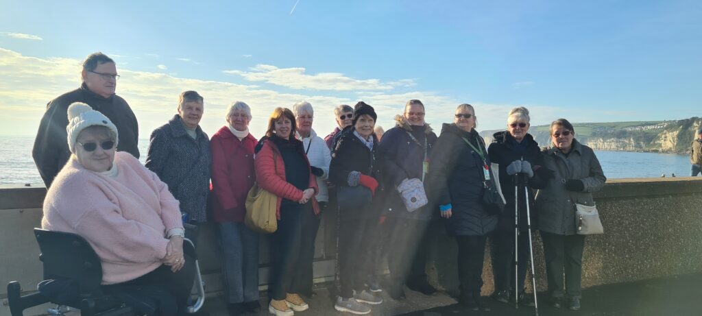 Group of Honitonn Carers at Seaton.. They are wearing winter clothes. The sea is in view behind them. 